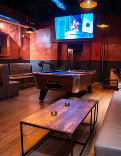 Industrial-style cannabis lounge showing a central pool table, television, exposed pipes, and rustic coffee tables with ashtrays in the foreground.