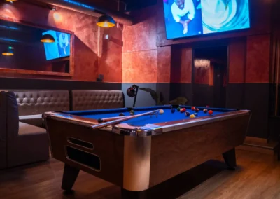Dark, moody lounge area featuring a pool table, large television displaying faces, and a wall mirror reflecting the ambient light.